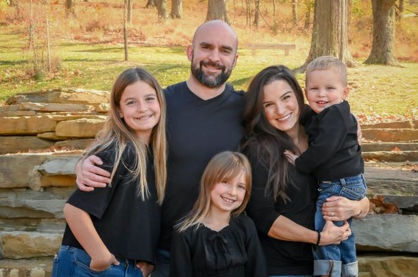 Family of five smiling outdoors on stone steps in a sunny park setting