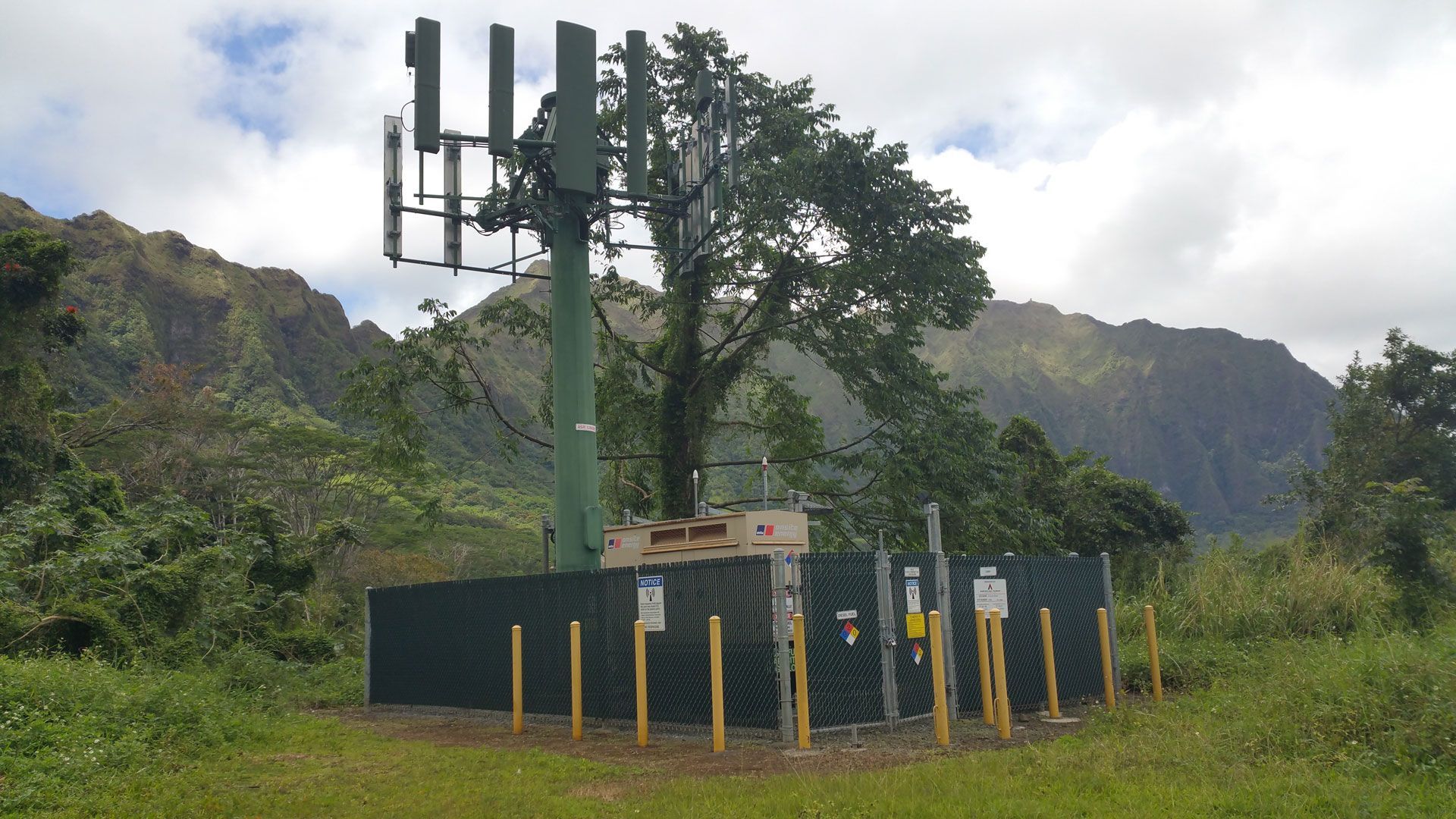 Cell tower surrounded by fenced equipment in a green, mountainous landscape.
