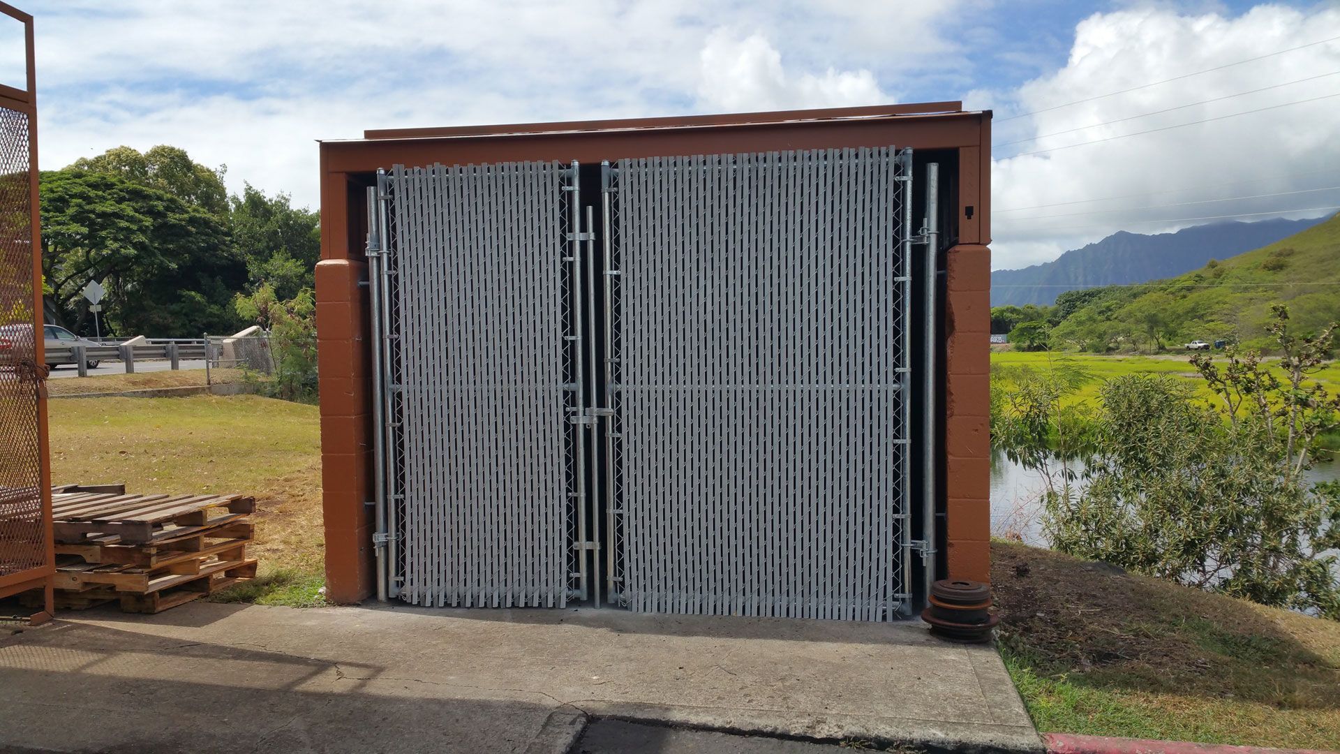 Large metal security gate, open. Brown frame with vertical bars, green foliage, mountains in the background.