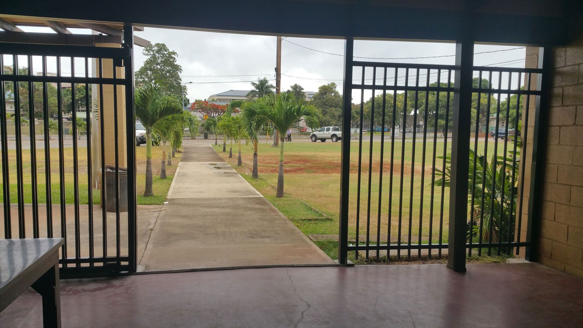 View through gate: pathway lined with trees, field, vehicles, and sky.
