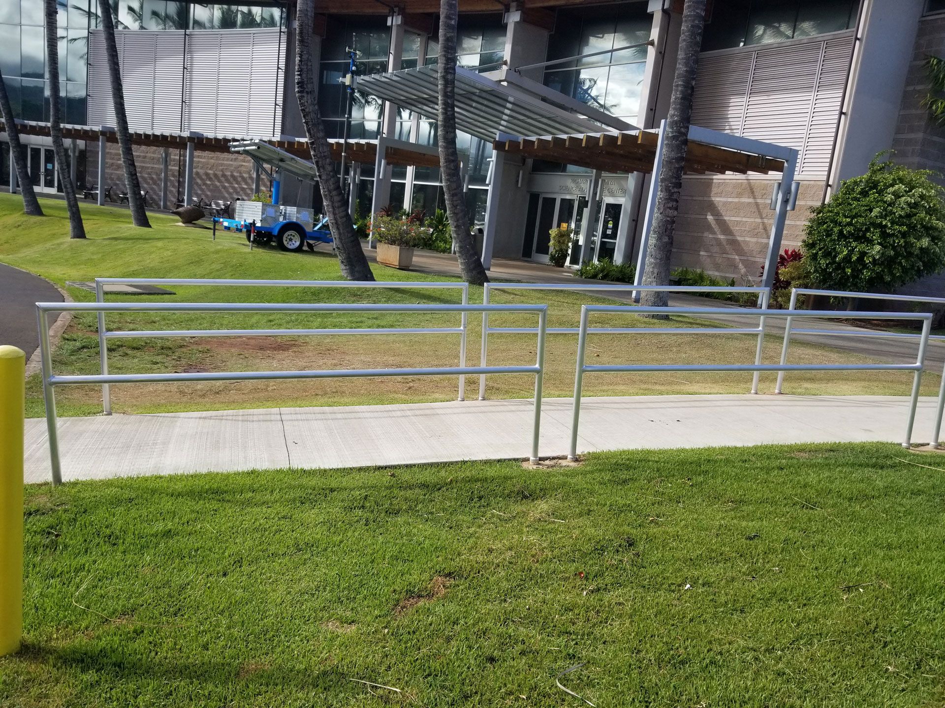 Railing along a sidewalk in front of a building with a canopy and glass doors; grass and trees are in the foreground.