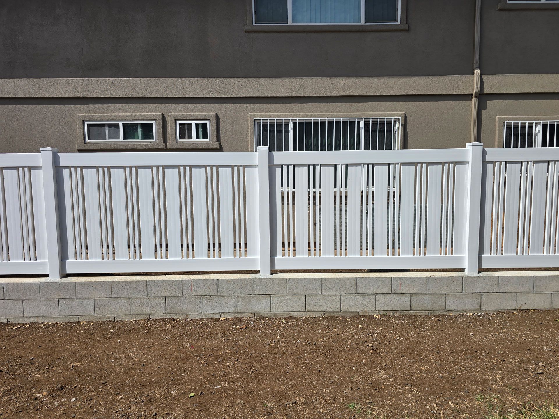 White vinyl fence with vertical slats atop a gray brick base. Brown dirt in front, building behind.