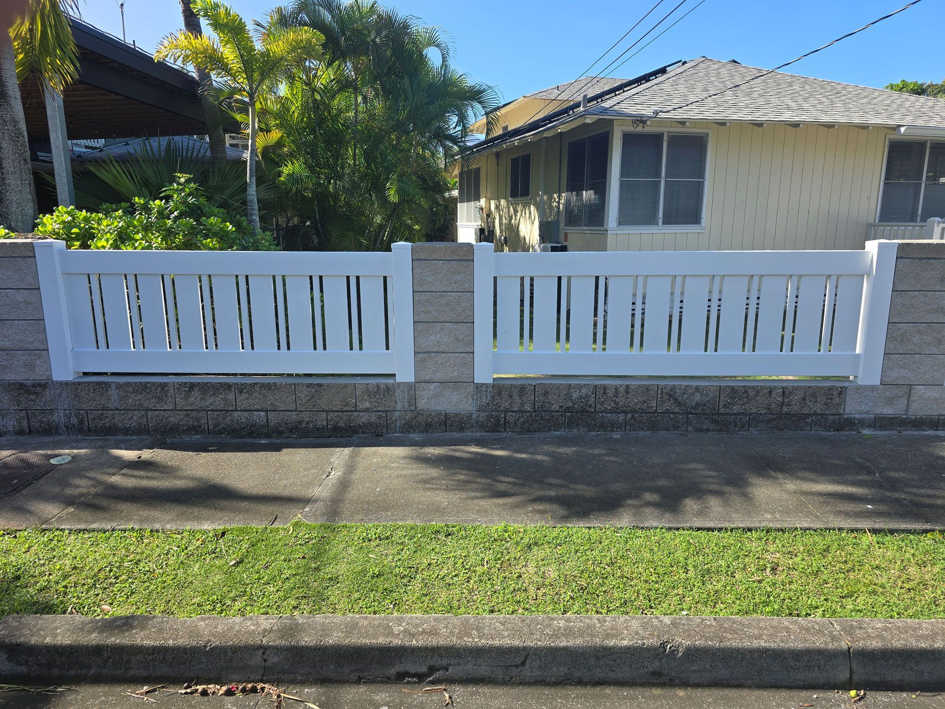 White picket fence between stone pillars, bordering a sidewalk and grass, with houses in the background.