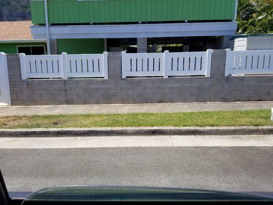 A white fence surrounds a brick wall in front of a green house.