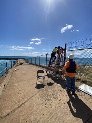 Two men are working on a chain link fence near the ocean.
