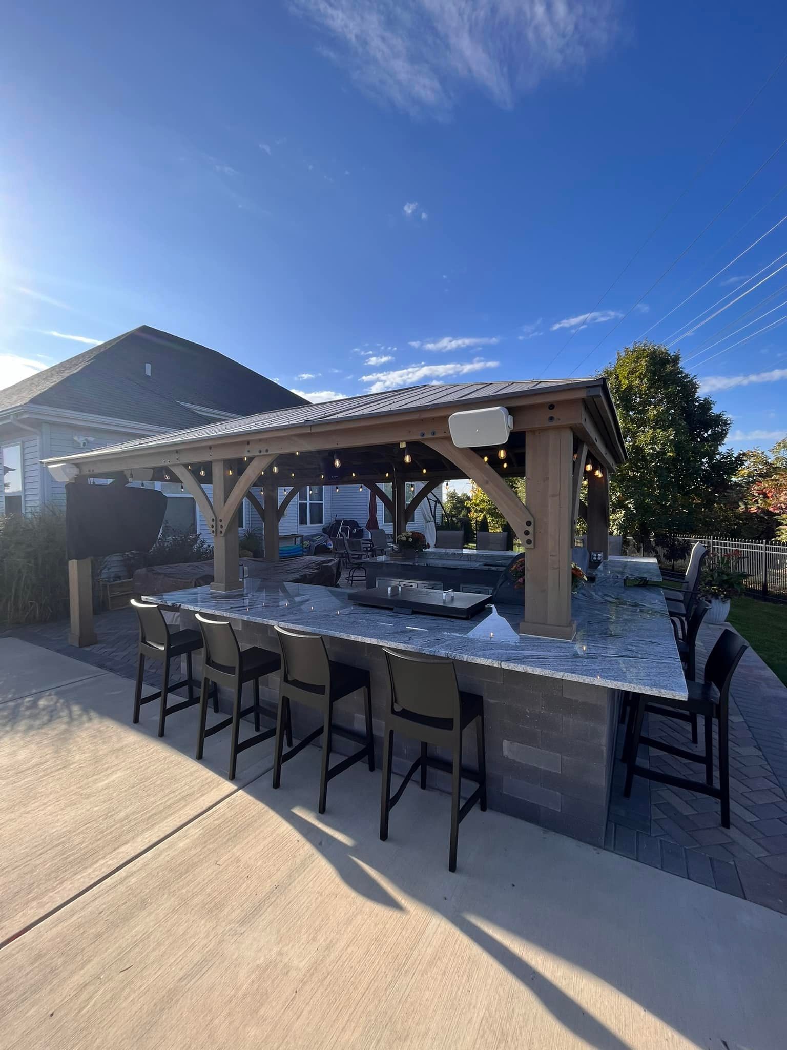 A wooden gazebo with a bar and stools in front of a house.