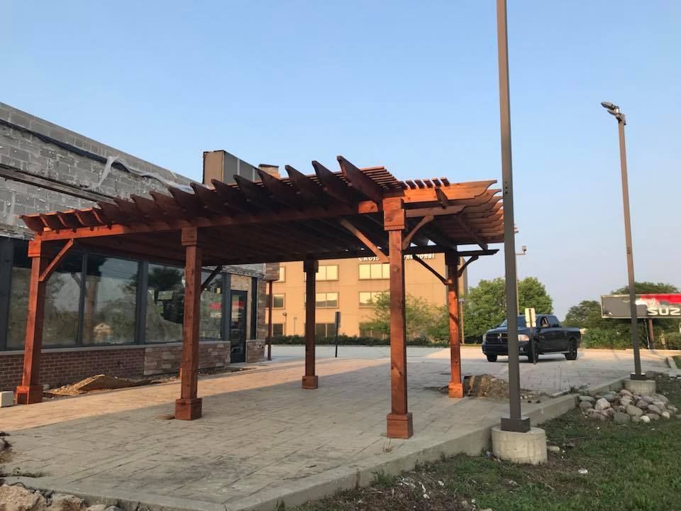 A car is parked under a wooden pergola in front of a building.