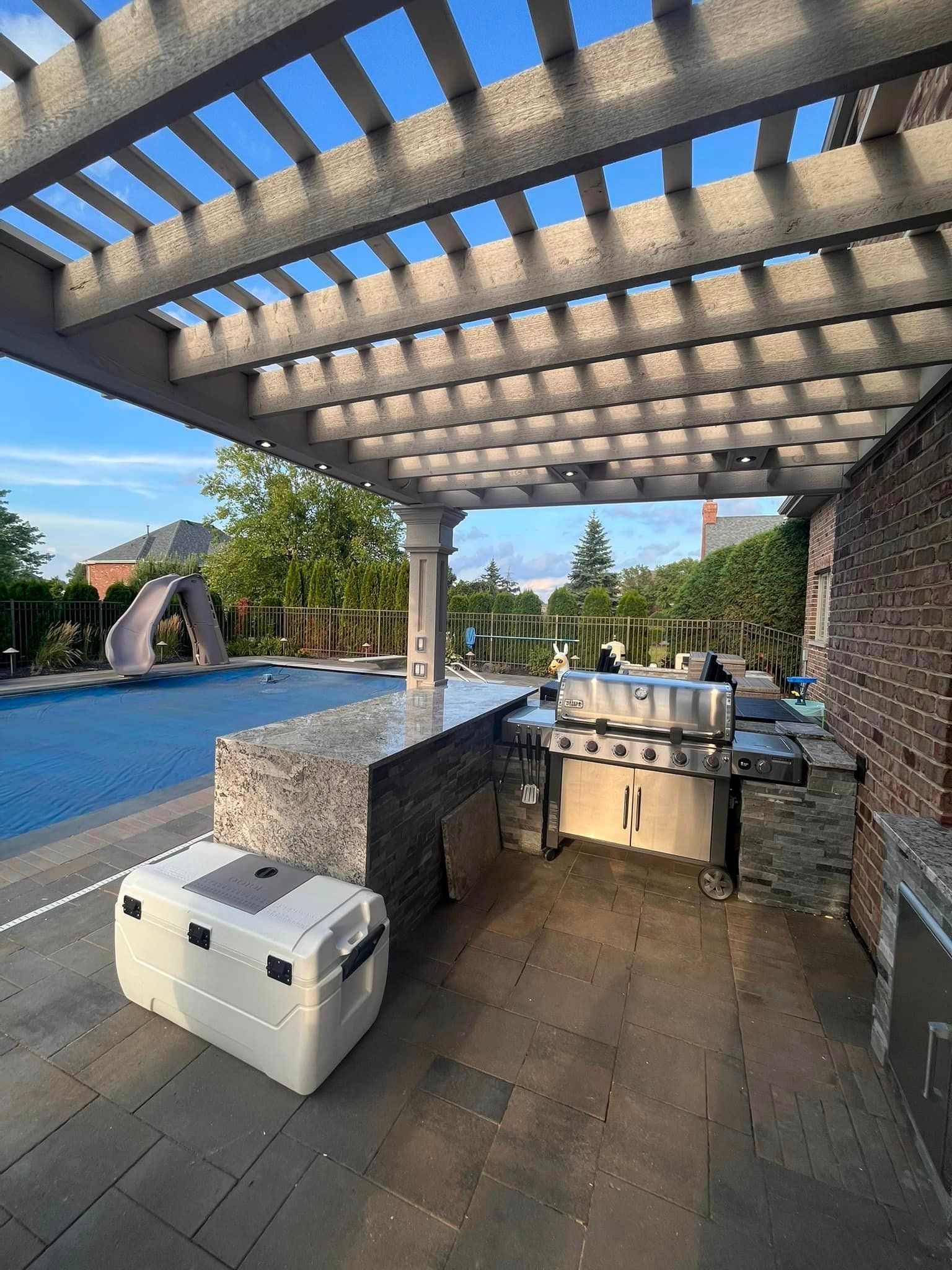 A large cooler is sitting under a pergola next to a swimming pool.