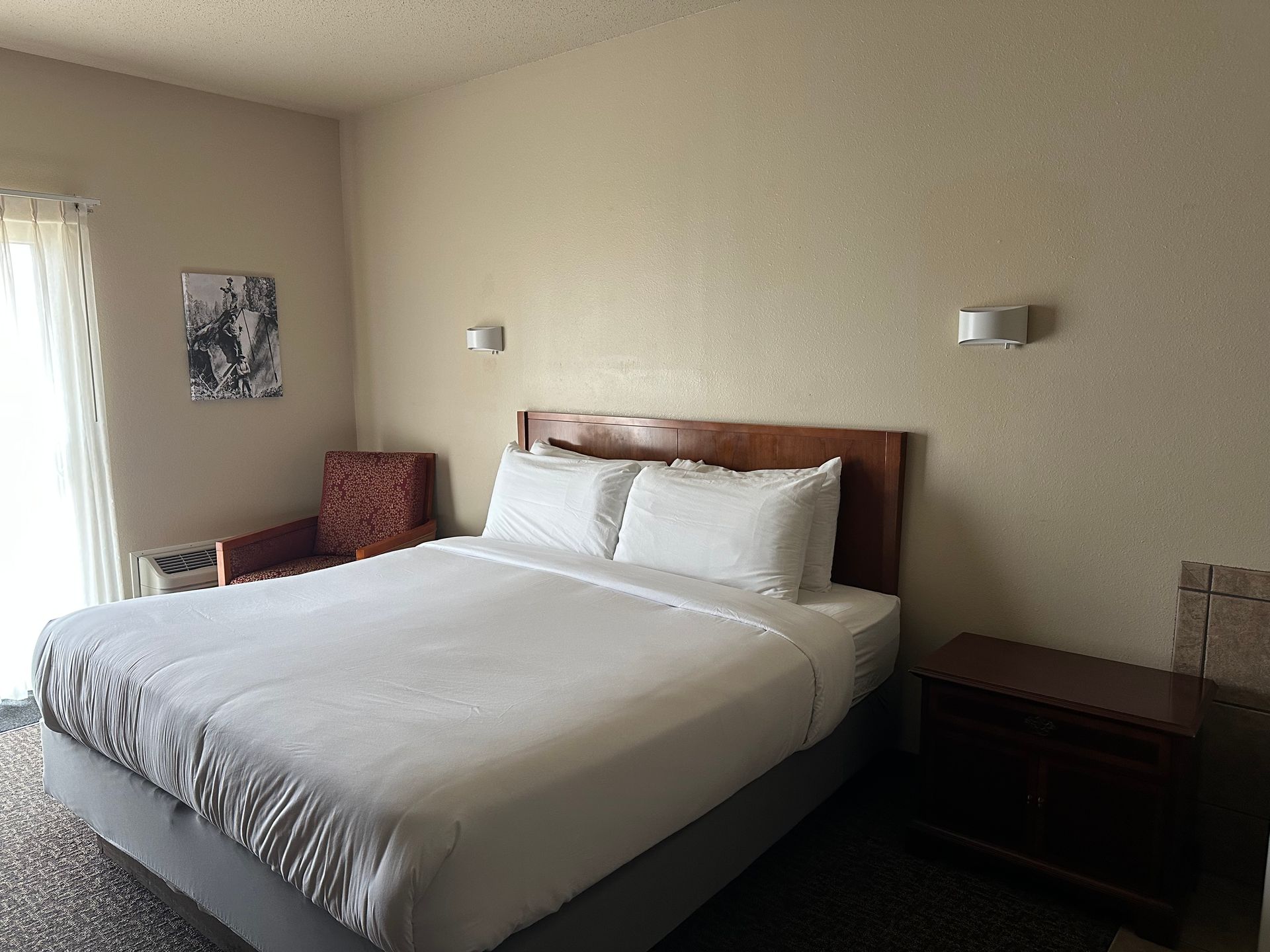 Bedroom with a bed, nightstand, chair, and artwork. Beige walls and a sliding glass door with curtains.