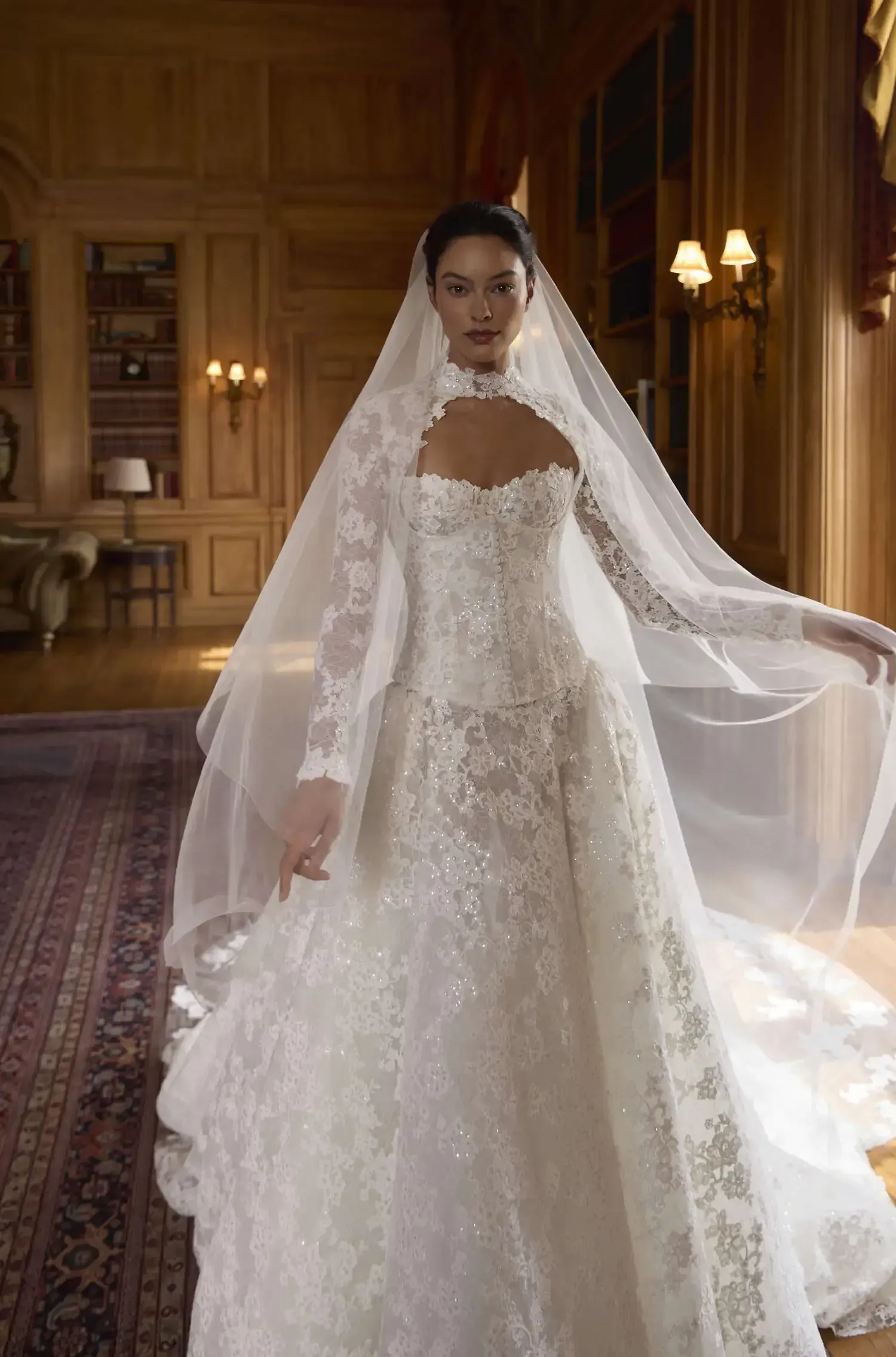 Bride in a white lace wedding gown and veil standing in an elegant room with chandeliers.