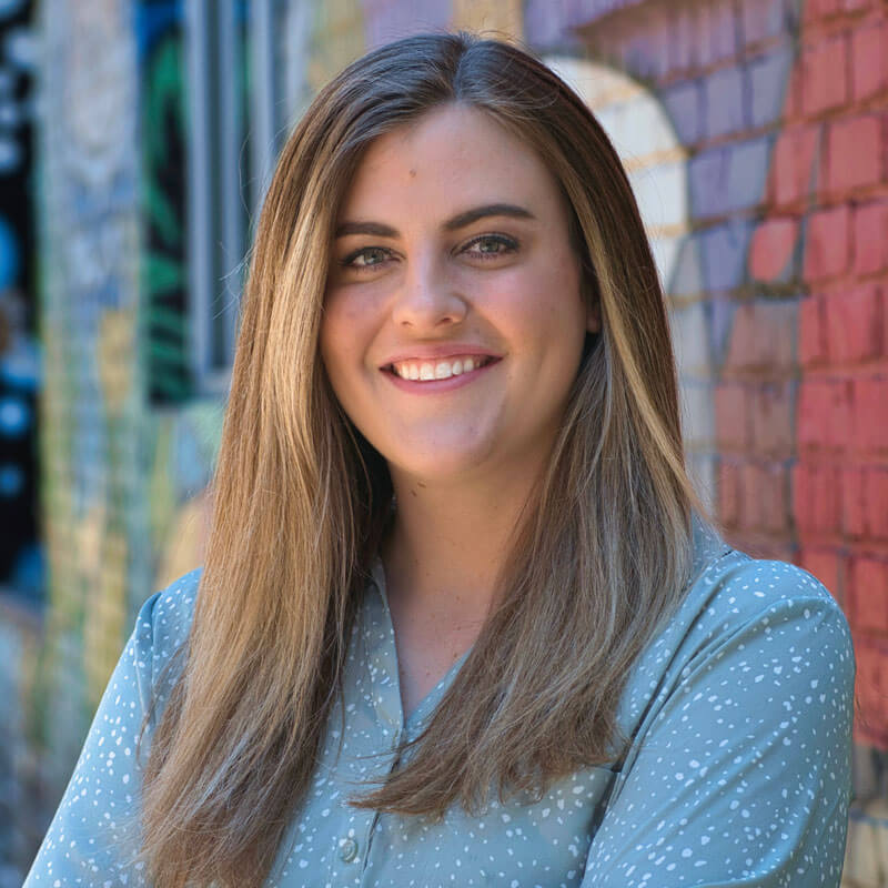 Woman with light brown hair smiles in front of a colorful brick wall. She wears a blue patterned top.