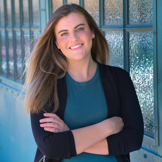 Woman with long hair smiles, arms crossed, wearing a dark jacket and teal shirt, standing in front of a blue window.