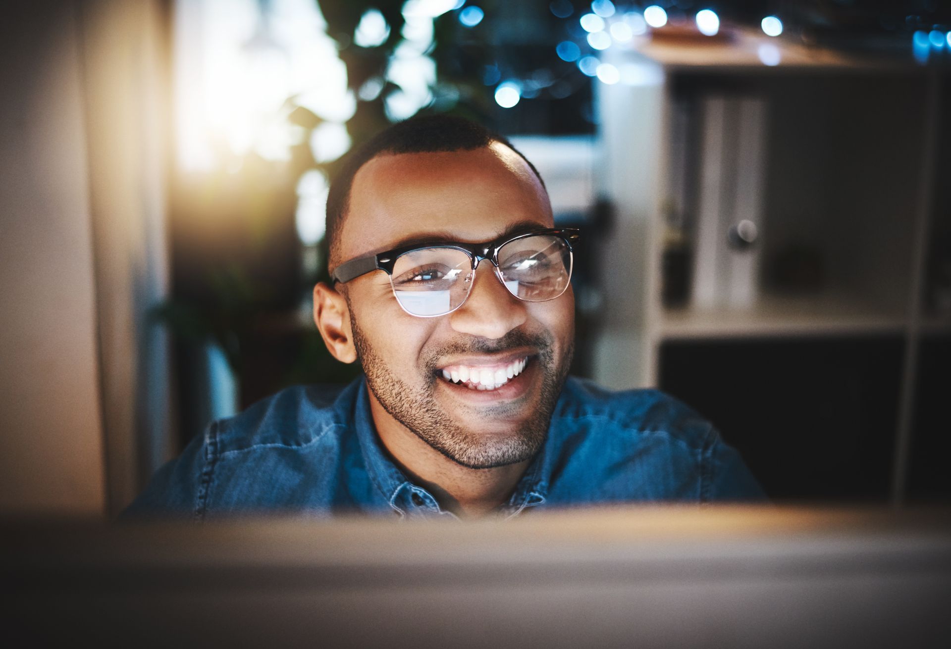 Man smiling while wearing glasses in a dim office setting, illuminated by a computer screen.