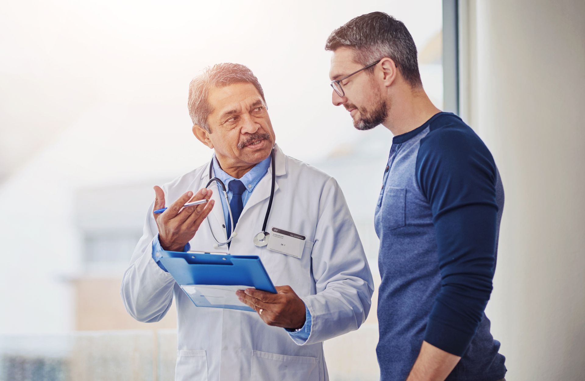 Doctor in lab coat discusses results with a patient, holding a clipboard, near a window.