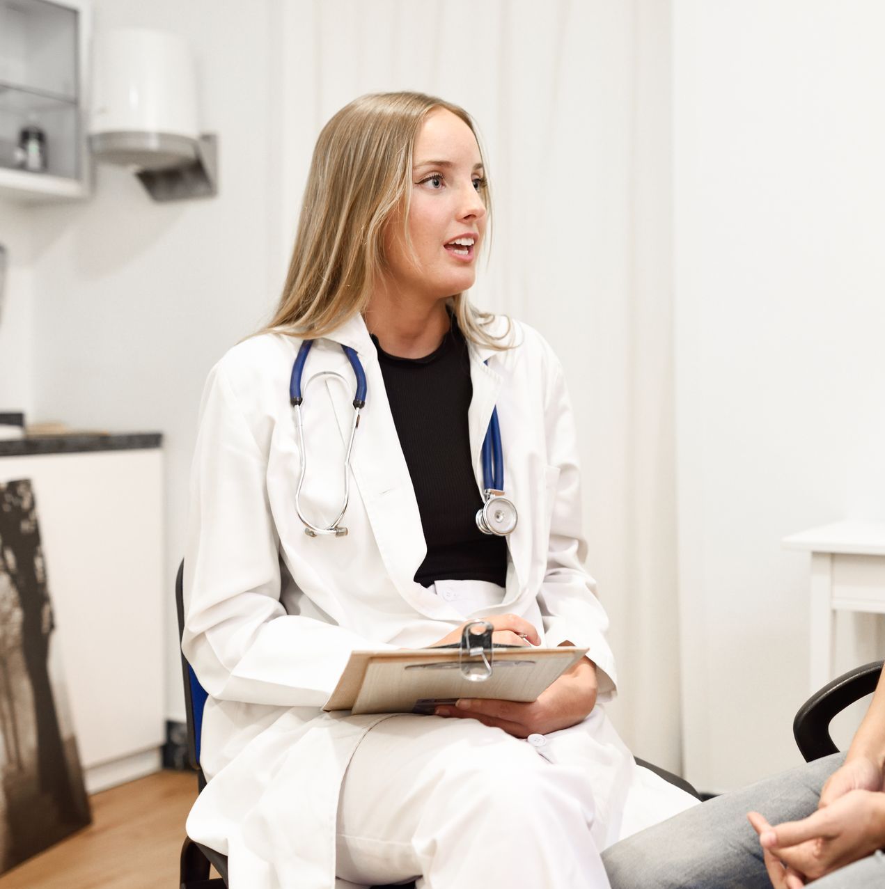 Doctor in a white coat with a stethoscope around her neck, holding a clipboard, talking to a patient in a medical office.