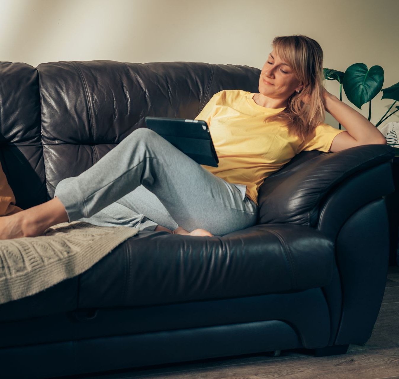 Woman on a black leather couch, reading a tablet. She wears a yellow shirt and gray pants in a room with a plant.