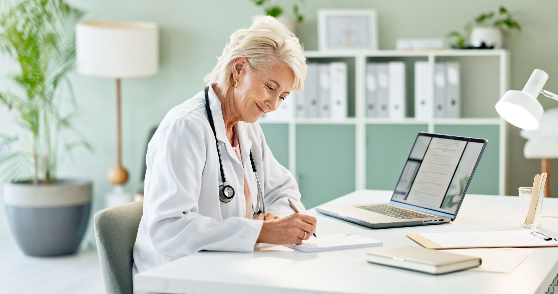 Doctor in white coat, stethoscope, writing at desk with laptop in office, smiling.