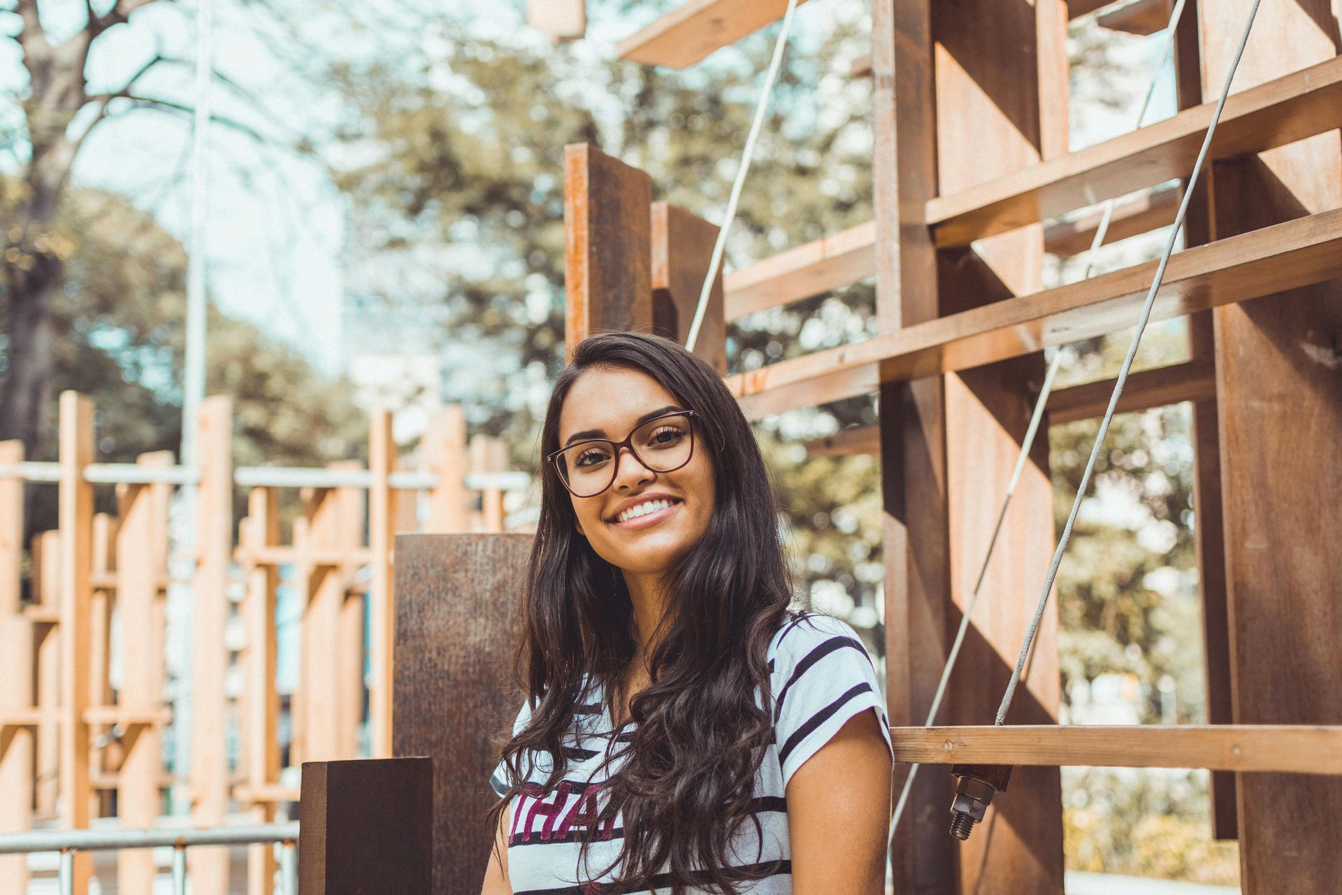 Woman with glasses smiles in front of a wooden structure outdoors.