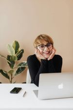 Woman smiling, wearing glasses, at a white desk with laptop, phone, pen, and plant.