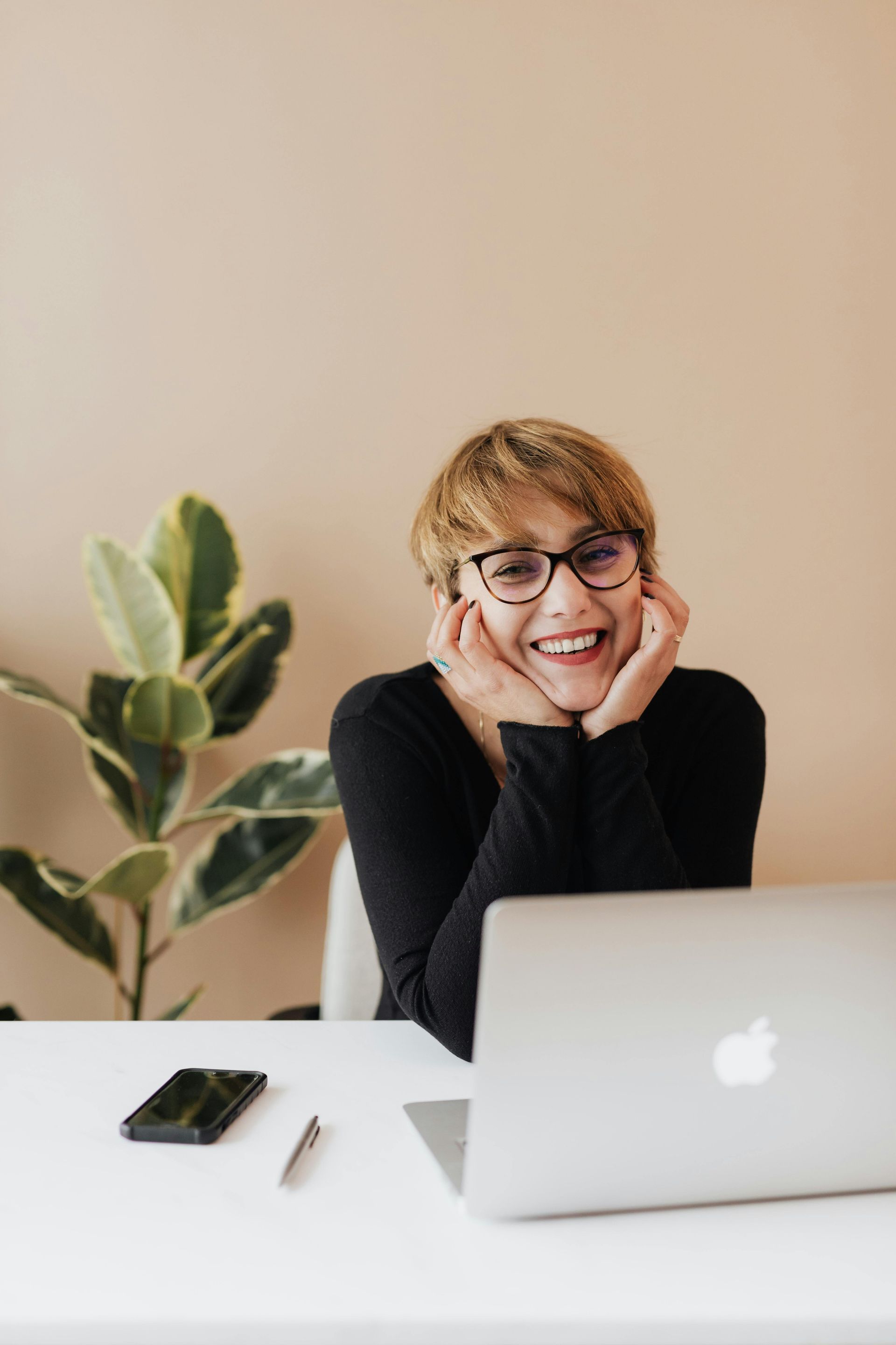 Woman smiling, wearing glasses, at a white desk with laptop, phone, pen, and plant.