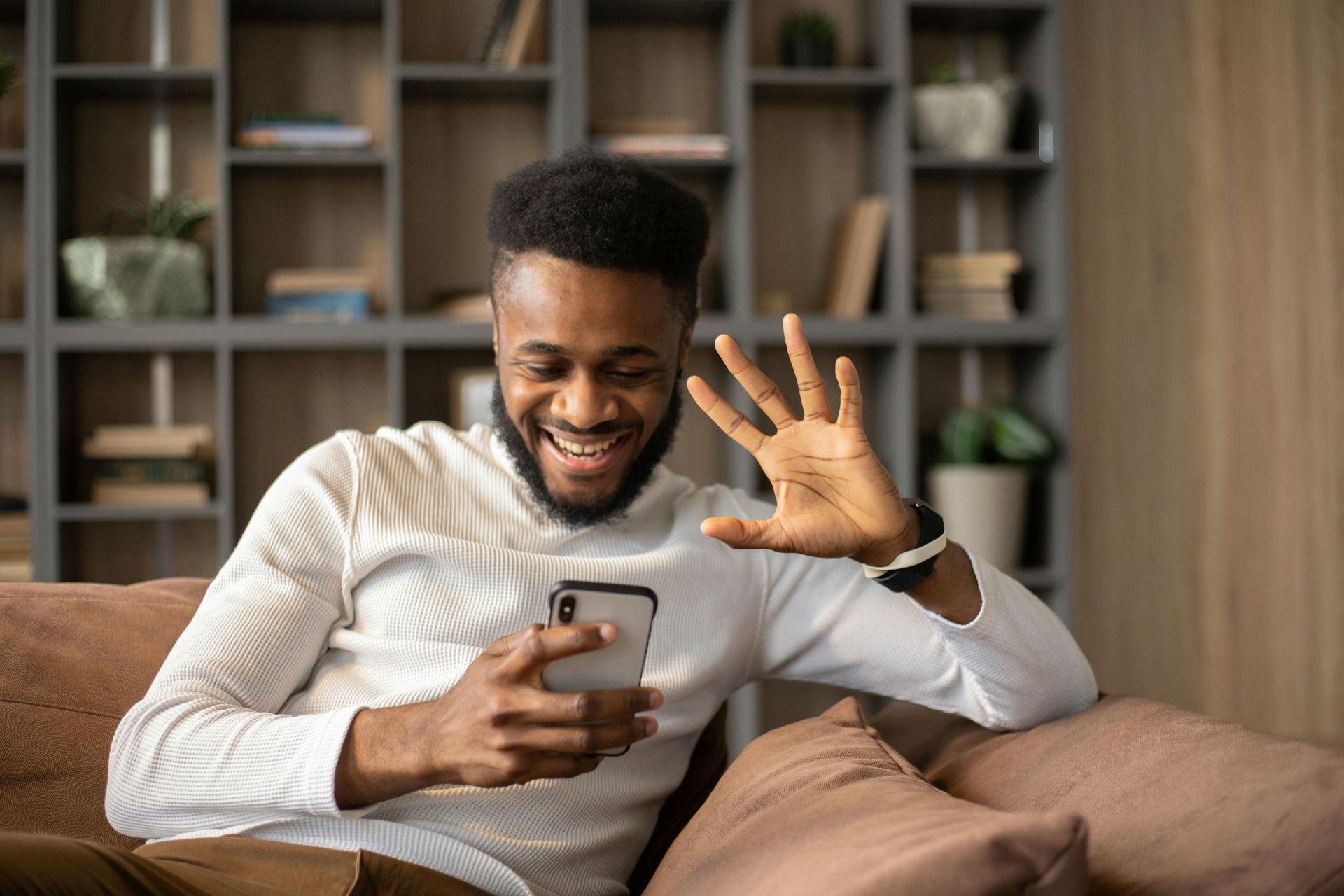 Man on a sofa smiles and waves at a phone; wood shelves in background.