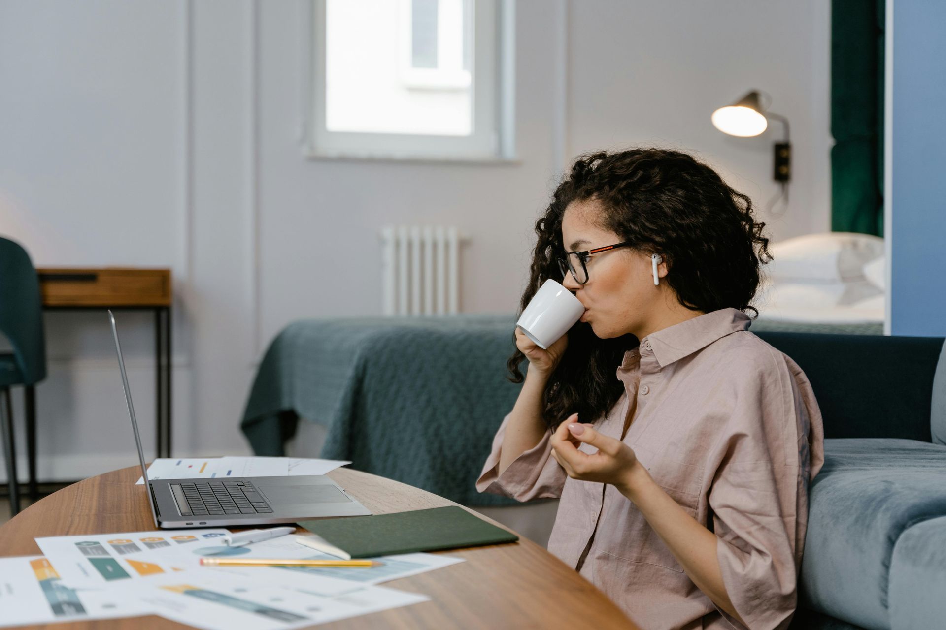 Woman drinks coffee while working on laptop at low table; documents spread around.