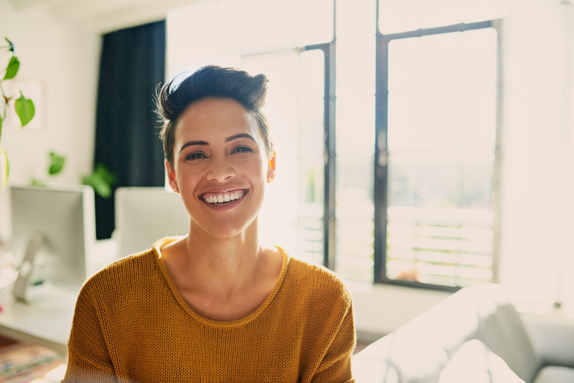 Woman smiling, indoors, sunlit, wearing a mustard-colored sweater, with short dark hair, desk and windows in the background.