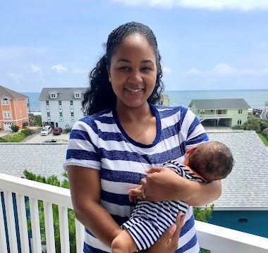 Woman in striped shirt holding baby on a balcony overlooking ocean and houses.
