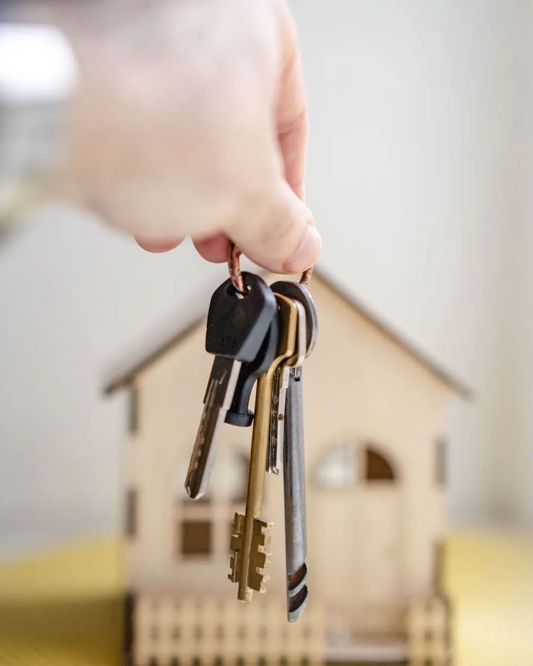 Hand holding a set of keys in front of a small wooden house model.