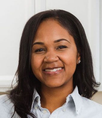 Woman smiling, wearing a light blue button-down shirt, in a well-lit indoor setting.