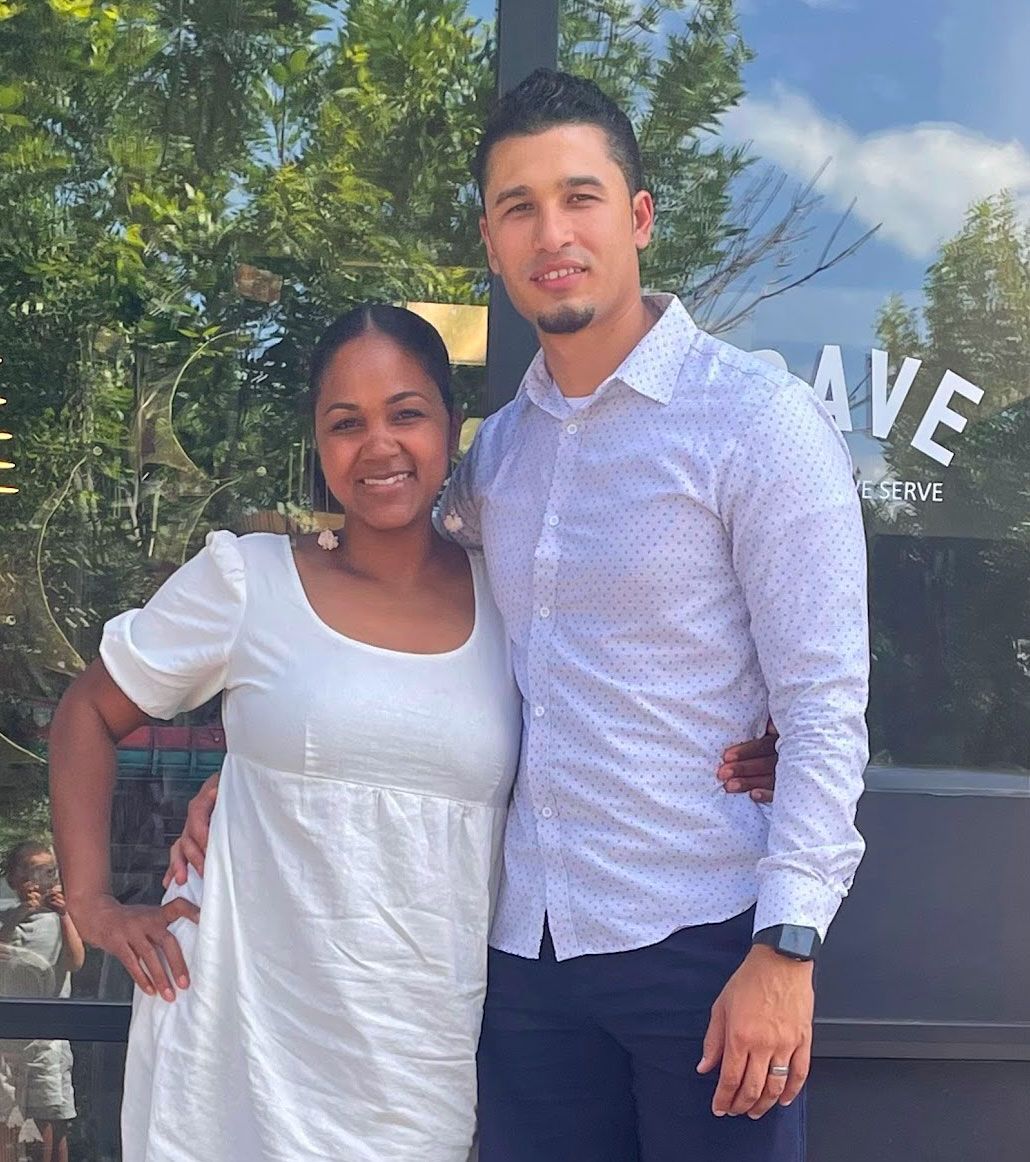 Couple smiling, standing by a glass door. Woman wears white dress, man a button-down shirt.