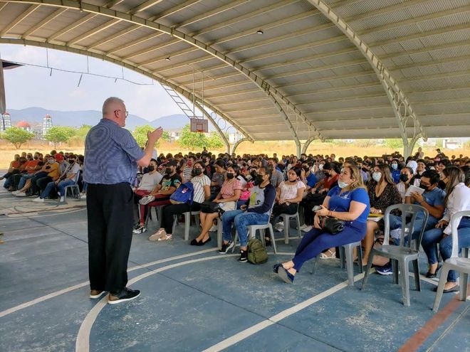 A man is standing in front of a crowd of people sitting in chairs under a canopy.