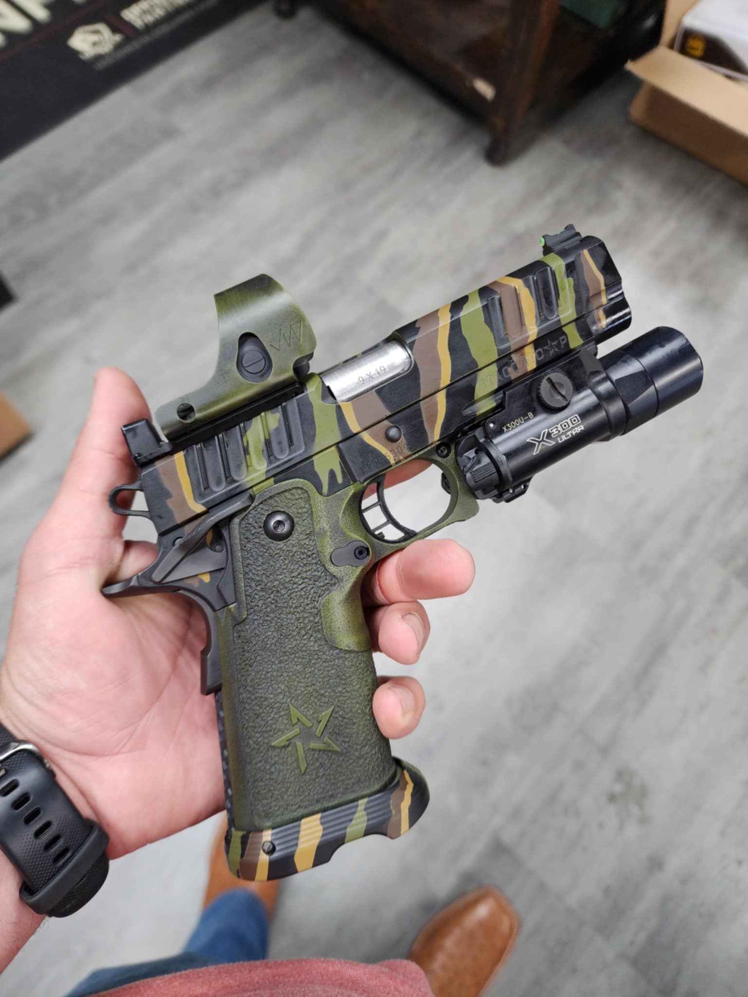 Hand holding a custom camouflage pistol with a red dot sight and flashlight.