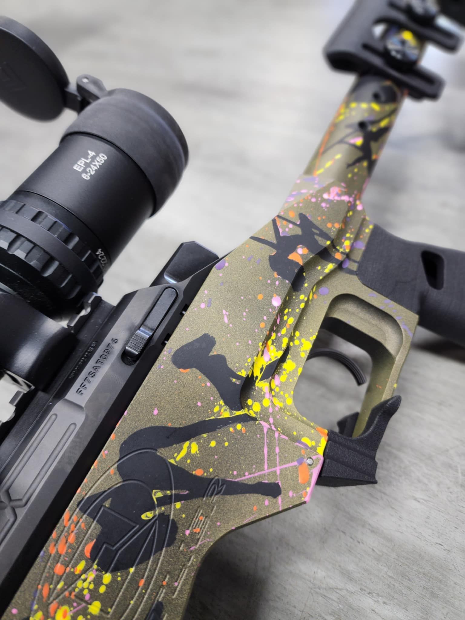 Custom-painted rifle with scope. Olive green base with splattered pink, yellow, and black design; indoor setting.
