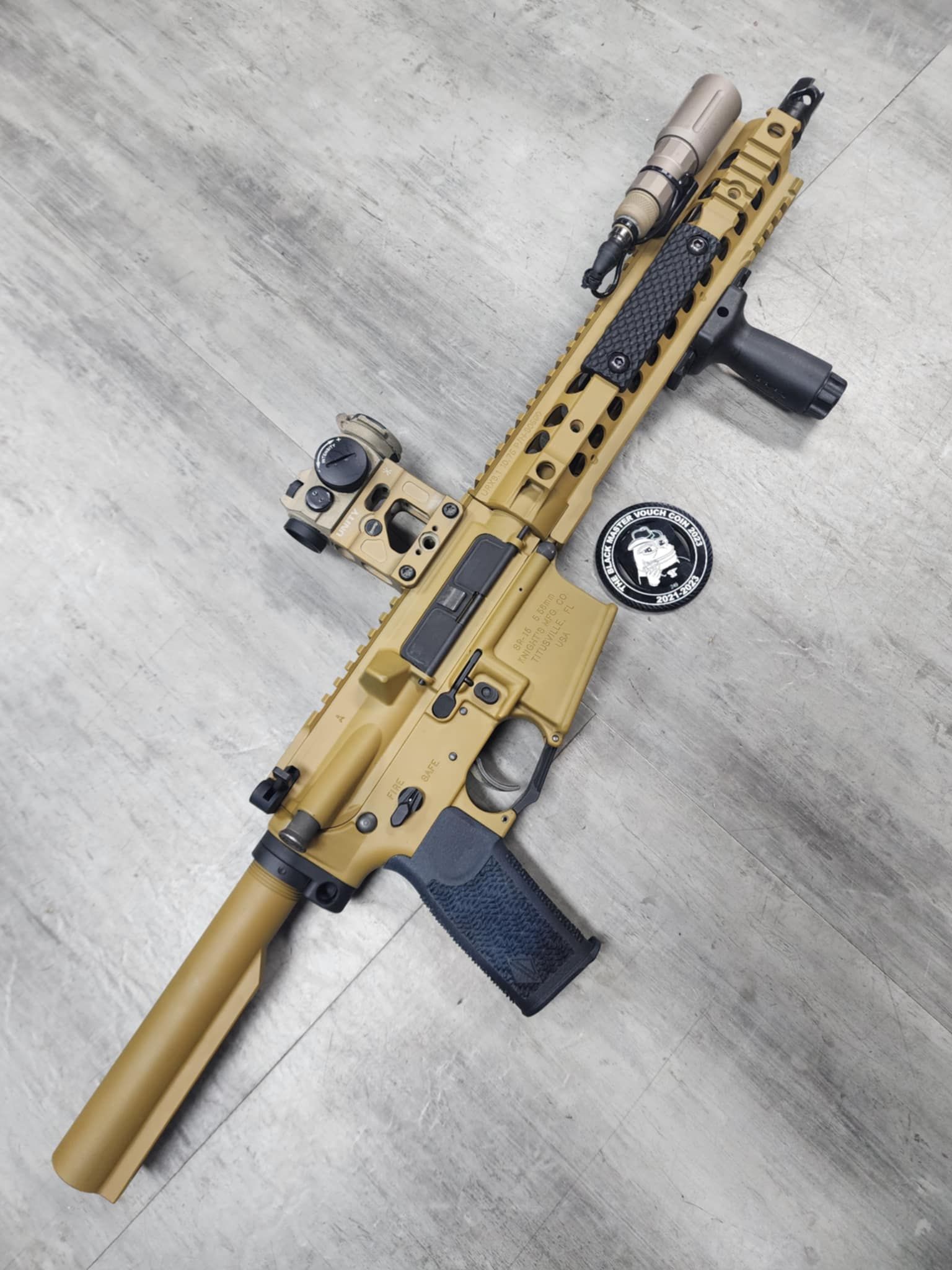 Tan and black AR-15 rifle with accessories, set on a gray wood floor.