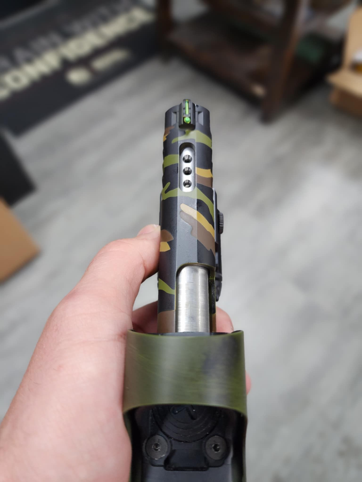 Hand holding a camouflage pistol, green front sight, with a black base.