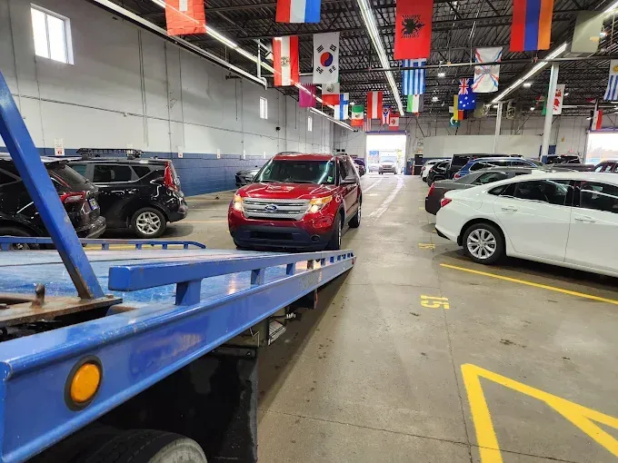A red SUV being unloaded from a blue tow truck inside a car dealership. Other vehicles and flags are present.