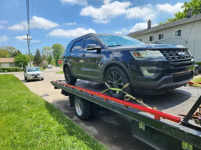 Dark SUV on a tow truck, secured by straps, parked on a residential street with a white car in the background.