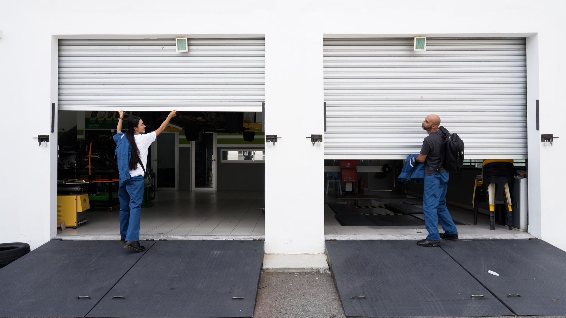 Two people, one opening a garage door, the other carrying bags. White building, gray doors.
