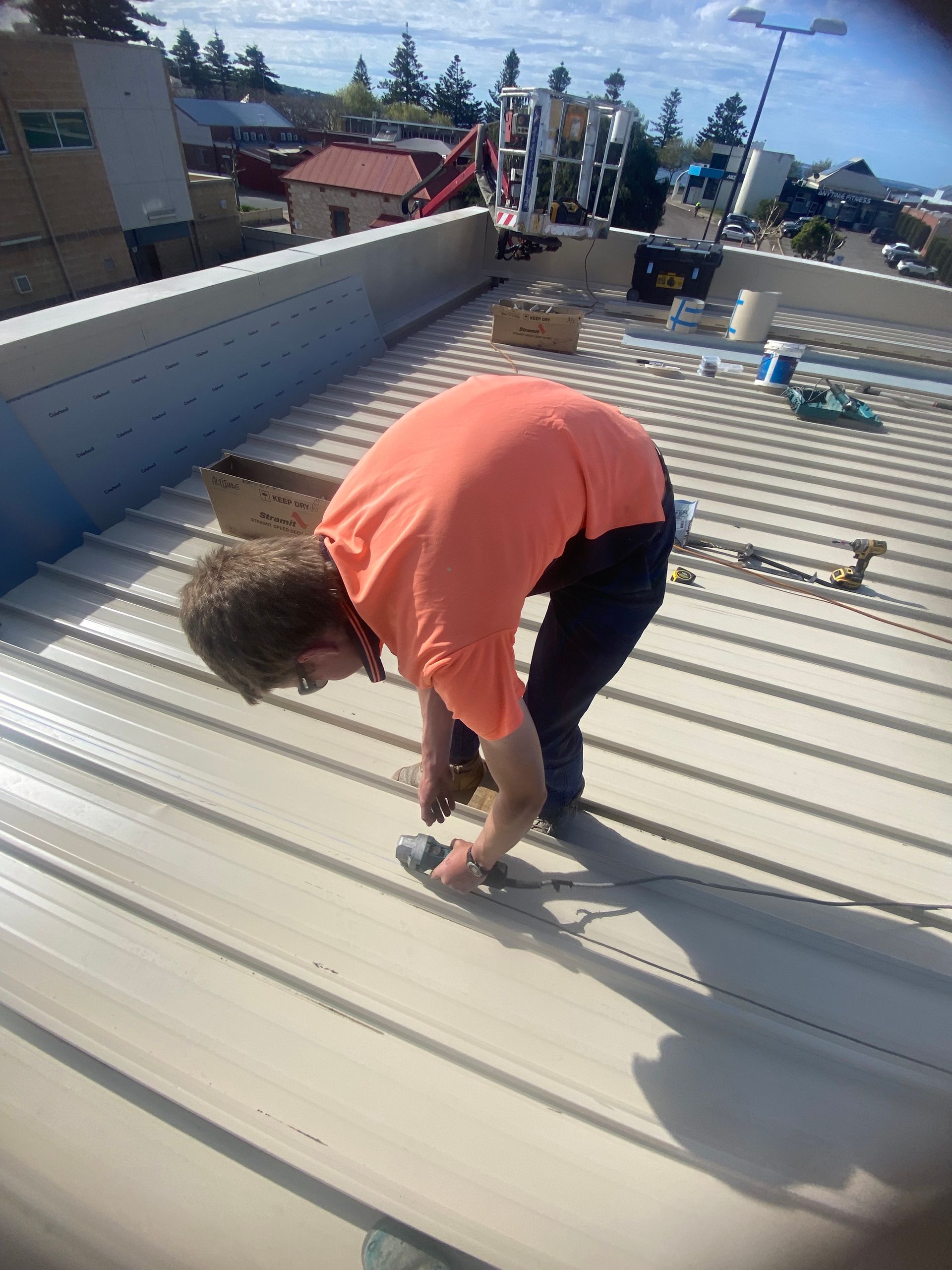 Person in orange shirt on a rooftop, working with a tool. Blue sky, industrial setting.