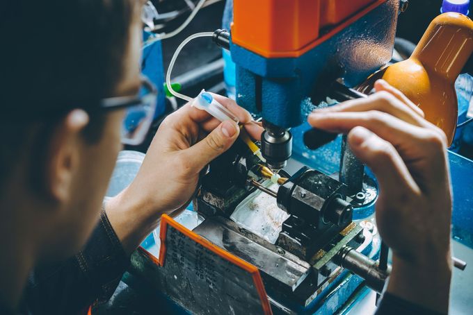 Primo piano, dall'alto, di una persona con occhiali di sicurezza che regola con precisione una piccola parte meccanica su un trapano a colonna.