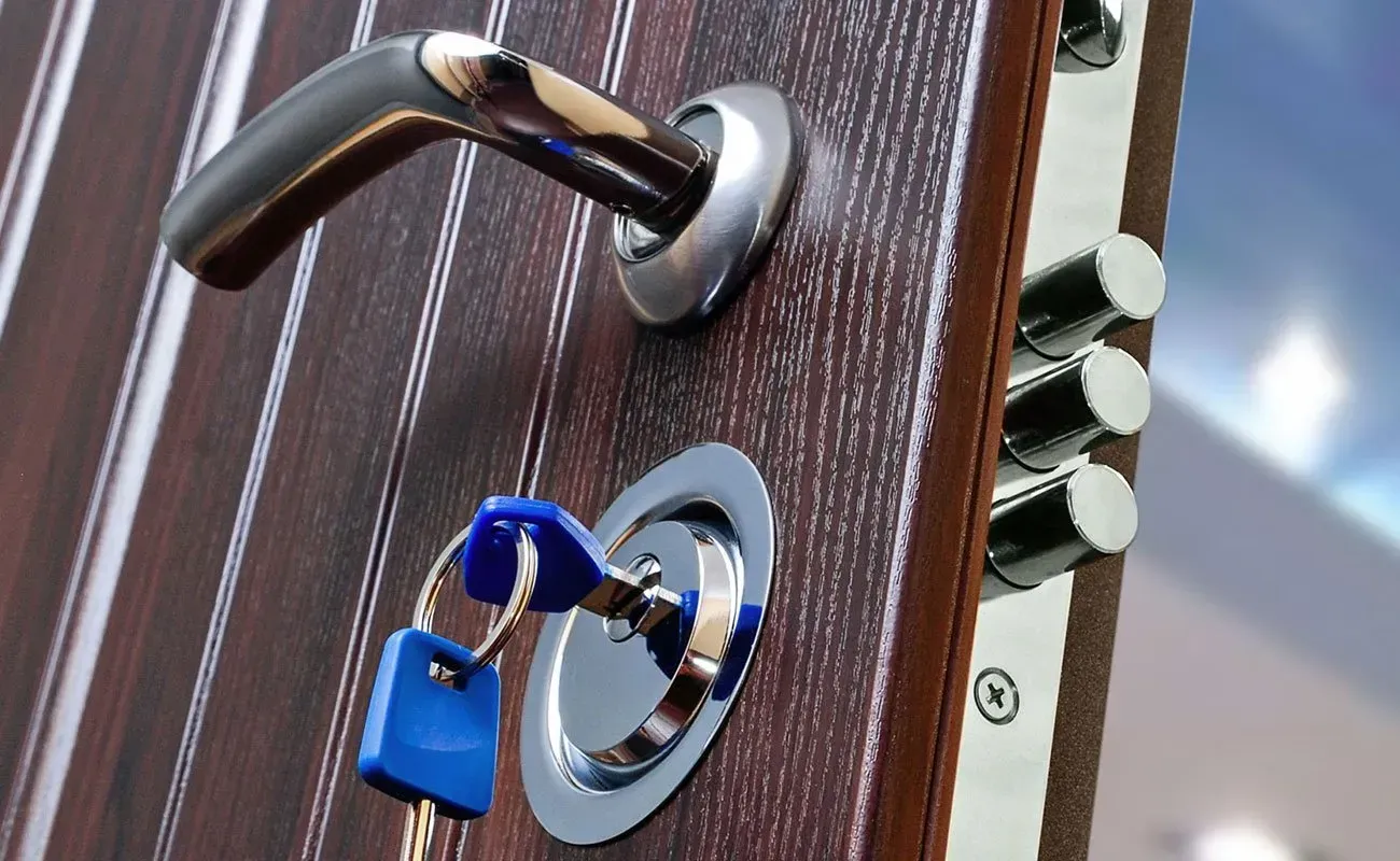 Primo piano di una porta con finitura effetto legno, maniglia cromata, chiave nella serratura e tre catenacci sporgenti.