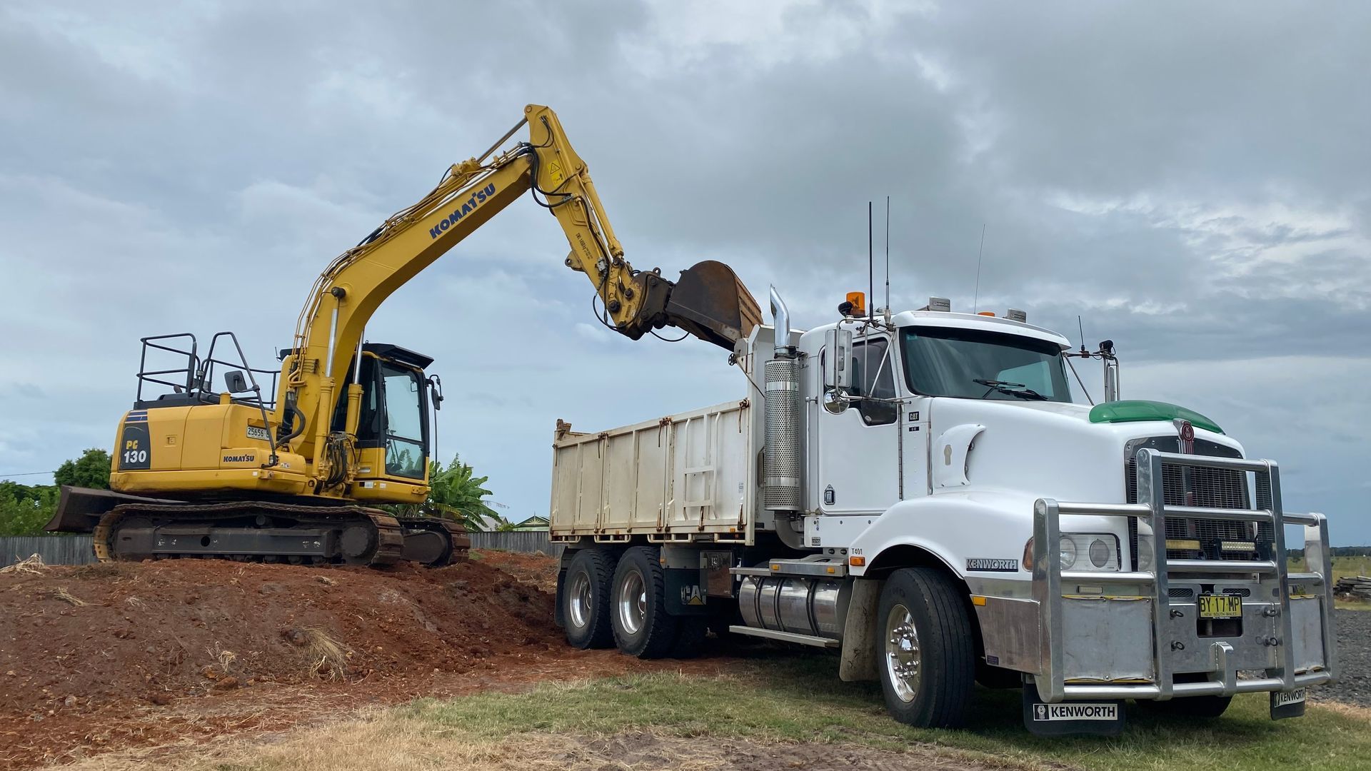 A Yellow Excavator Loading Dirt Into a White Dump Truck — PVM Contracting in Alstonville, NSW