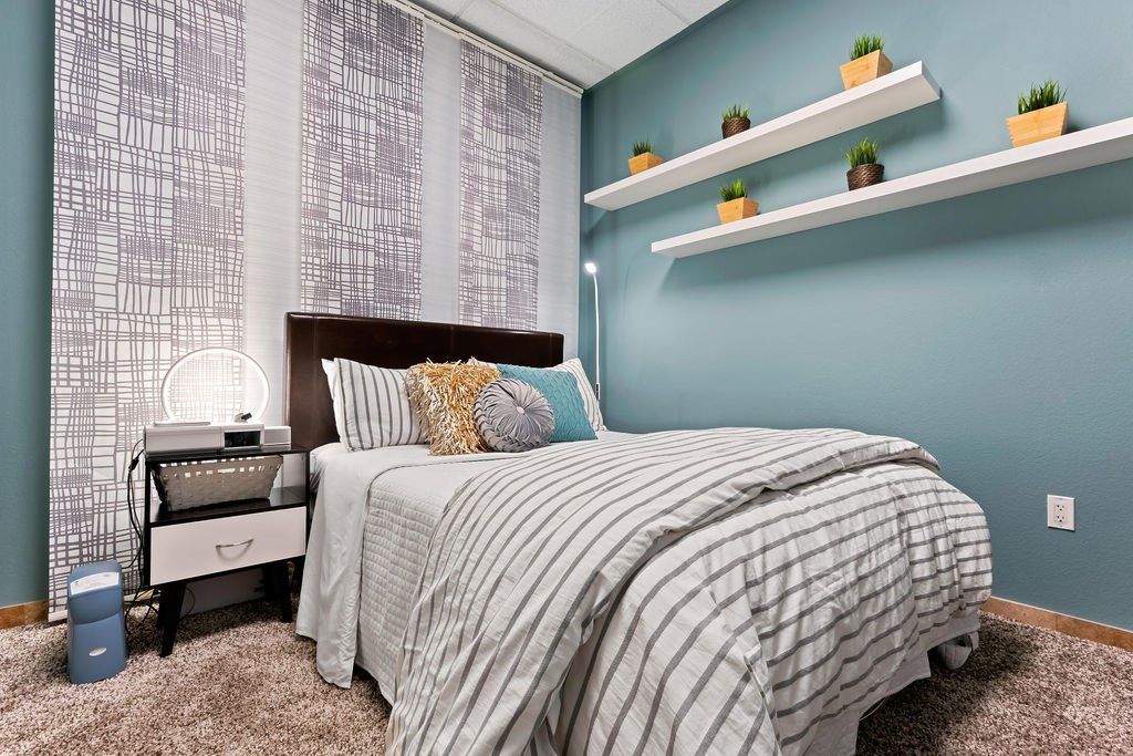 A bedroom with a bed , nightstand , shelves and a blue wall. | United Critical Care