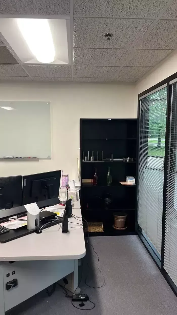 Small office with a desk, computer monitors, cabinet, whiteboard, and a glass door to the right.