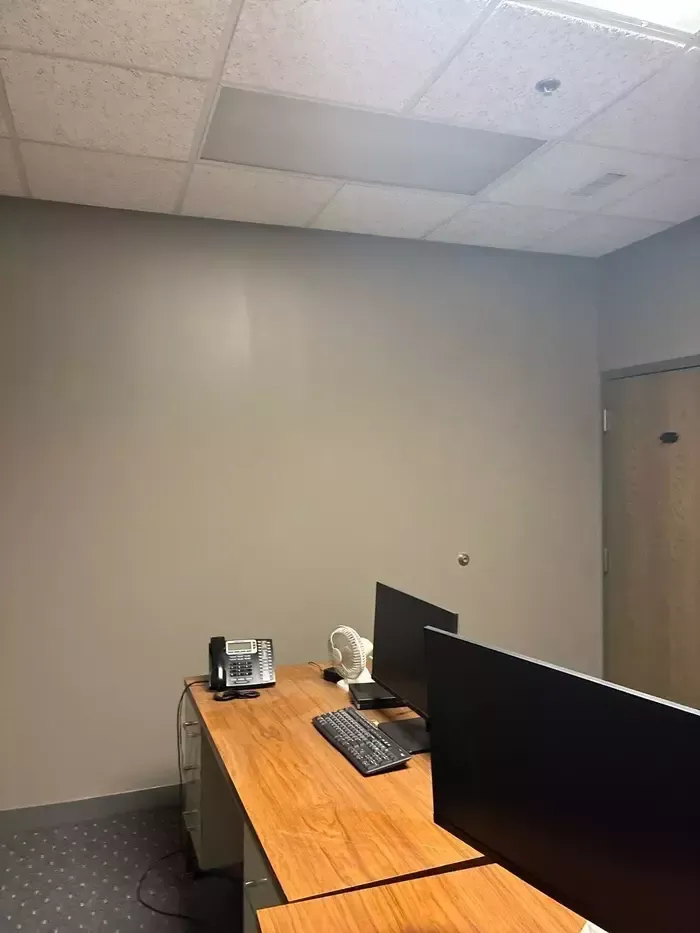 Small office with two computer monitors, keyboard, and wooden desks against a plain gray wall.