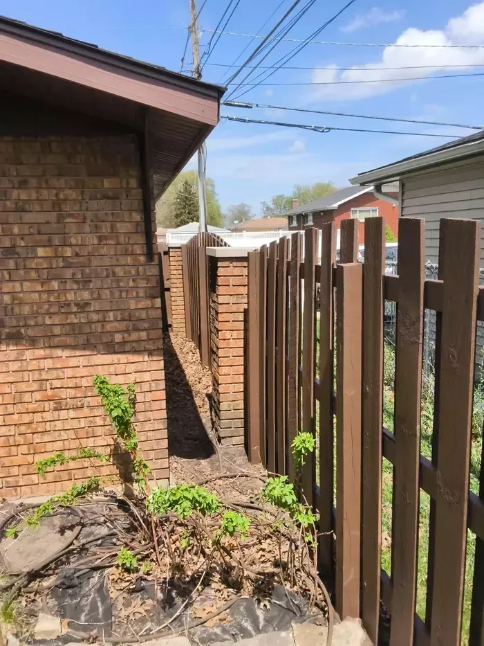 Narrow side yard between a brick house and wooden fence, with small plants and overhead utility lines.