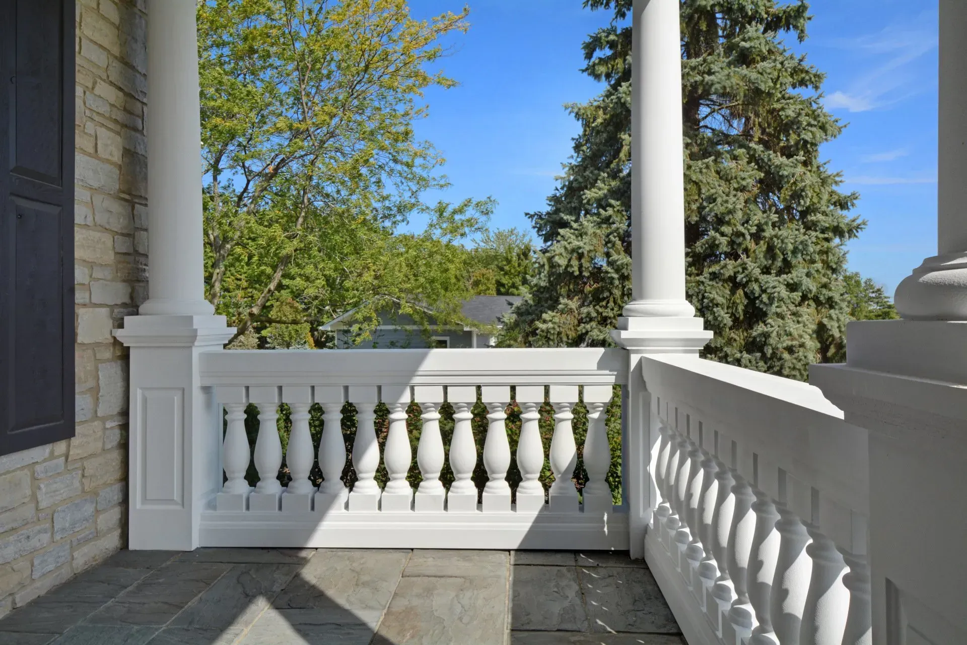 Front porch with white columns and balusters, stone floor, and a view of green trees under a blue sky.