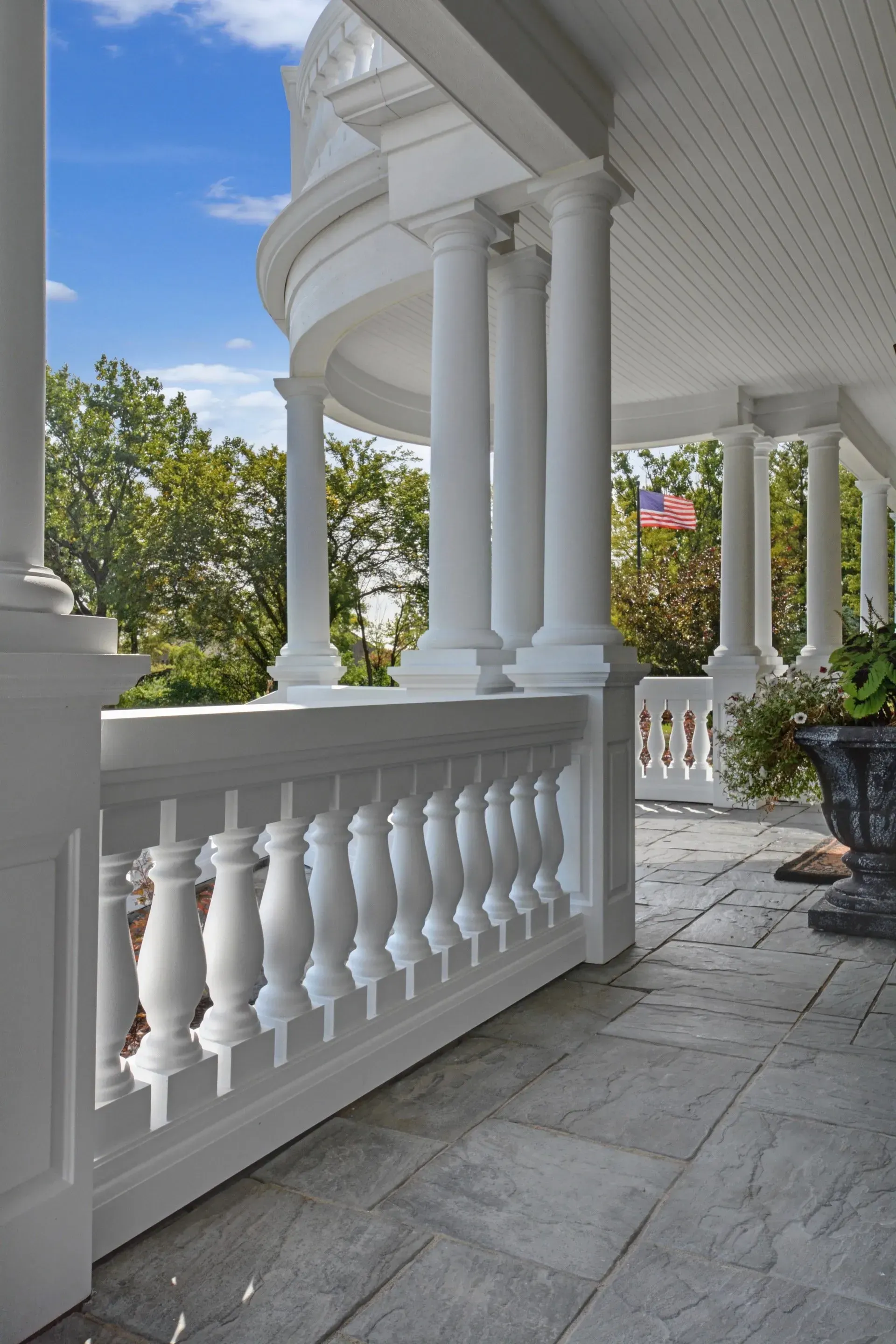White porch with columns, balustrade, and stone floor overlooking green trees and flowering bushes