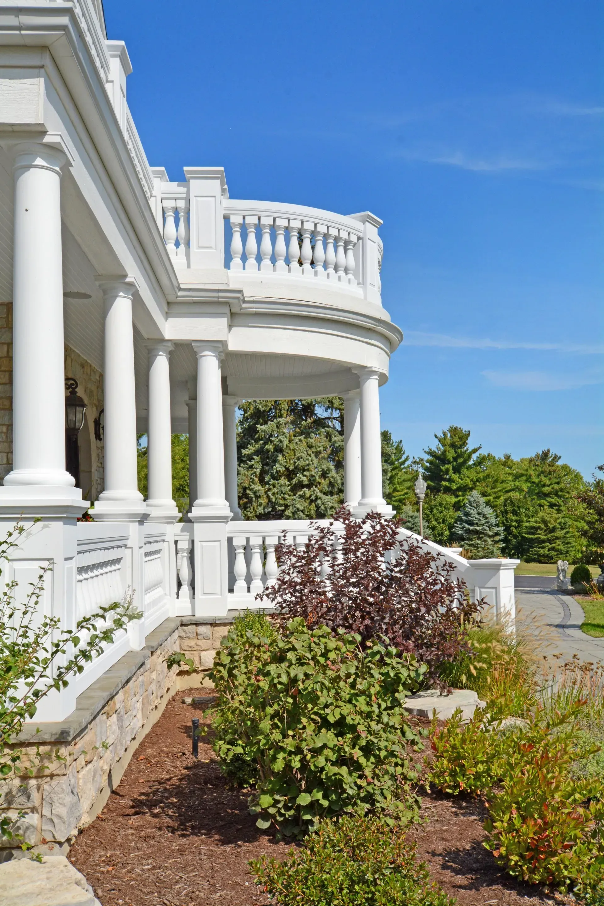White colonial-style porch with columns, balcony railing, and landscaped shrubs under a blue sky